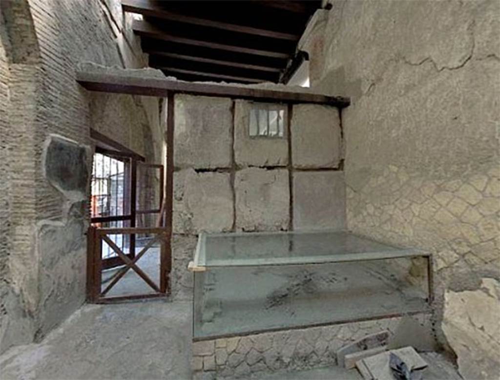 VI.21 Herculaneum. May 2006. Looking north across “caretaker’s room”, with remains of skeleton on its bed under a glass case.
Deiss wrote that this room had a door and a small rectangular window.
The window is placed high and is barred. In the room was a wooden table of good quality and an elegant wooden bed. On the bed is the skeleton of a man who had thrown himself face downwards in defence against the volcanic gases……. The supposition has been advanced that the little room was occupied by a caretaker or priest, and perhaps due to illness, he was unable to escape.
If the man was a caretaker, why should he have a temporary room adjoining the shrine? Why furniture of such high quality? If a priest, why not a room of more impressive construction? Why would the chief server at an emperor’s shrine be left behind on the flight of his fellows? And if these living quarters, in spite of their hurried construction, were intended to be permanent, why no stove, no brazier for winter, no latrine? Neither hypothesis takes account of the barred window or the evident despair in the man’s position. It seems apparent that he could not escape, and no one unlocked the door……. Whatever the reason for the man in the barred room of the Augustales, in the end he too paid with his life. Perhaps someday a chance reference in literature will provide the key to that strange little room and its hopeless occupant.
See Deiss, J.J. 1968. Herculaneum: a city returns to the sun. UK, The History Book Club, (p.155).